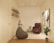 A cozy, minimalist room with neutral tones featuring a comfortable armchair, a bean bag, a small side table with plants, and decorative artwork on the walls. The shelves on the left hold various books and small decor items. The room has a modern white textured wall and wooden flooring with a geometric pattern rug.