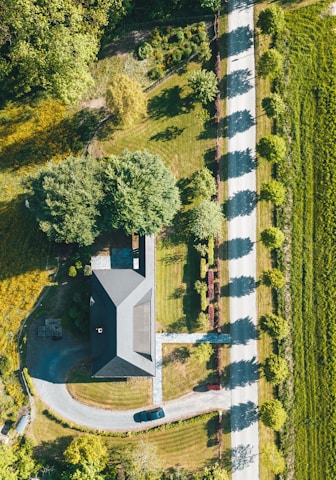 Aerial view of a house surrounded by a well-maintained lawn and trees, with a car parked on a curved driveway. The property is bordered by a straight road on the right, with shadows cast by a row of trees lining the road. The landscape includes various greenery and appears to be in a rural or suburban area.