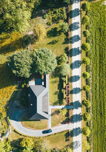 Aerial view of a house surrounded by a well-maintained lawn and trees, with a car parked on a curved driveway. The property is bordered by a straight road on the right, with shadows cast by a row of trees lining the road. The landscape includes various greenery and appears to be in a rural or suburban area.