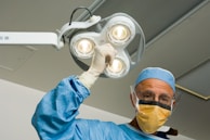 A surgeon wearing a blue surgical gown, cap, and gloves The surgeon is also wearing a protective face mask and glasses. The background appears to be an operating room with bright overhead lighting.