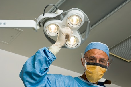 A surgeon wearing a blue surgical gown, cap, and gloves is looking directly at the camera while adjusting a ceiling-mounted surgical lamp. The surgeon is also wearing a protective face mask and glasses. The background appears to be an operating room with bright overhead lighting.