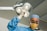 A surgeon wearing a blue surgical gown, cap, and gloves is looking directly at the camera while adjusting a ceiling-mounted surgical lamp. The surgeon is also wearing a protective face mask and glasses. The background appears to be an operating room with bright overhead lighting.