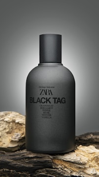 A sleek, matte black bottle with a textured cap is prominently displayed. The label reads 'ZARA BLACK TAG', with notes of grapefruit, bergamot, pepper, musk, vetiver, and vanilla listed below. The bottle rests on textured wood, providing an earthy contrast to the smooth surface of the bottle.