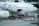 The image depicts an airplane on the tarmac at an airport with ground crew members working around it. The aircraft has elaborate black and white illustrations of animated characters on the fuselage. Cargo or luggage is being loaded into the airplane via specialized equipment, while ground personnel wearing safety gear are managing the process.
