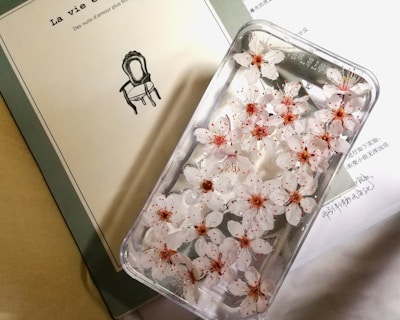 A transparent phone case is filled with delicate white flowers with pink centers, placed on top of a light-colored book and a sheet of paper with printed text. The flowers are carefully arranged, showcasing their intricate petals and adding a touch of natural beauty to the objects they rest on.