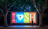 A brightly lit billboard in the evening featuring the text 'I ❤ SH' in large letters. The billboard is divided into sections with a heart symbol in the middle, and there is a person walking by in silhouette. Two large trees frame the billboard on either side, with dark foliage in the background.