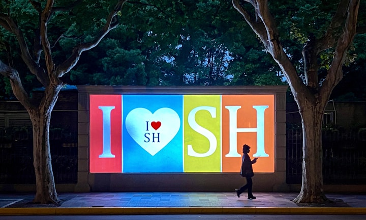 A brightly lit billboard in the evening featuring the text 'I ❤ SH' in large letters. The billboard is divided into sections with a heart symbol in the middle, and there is a person walking by in silhouette. Two large trees frame the billboard on either side, with dark foliage in the background.