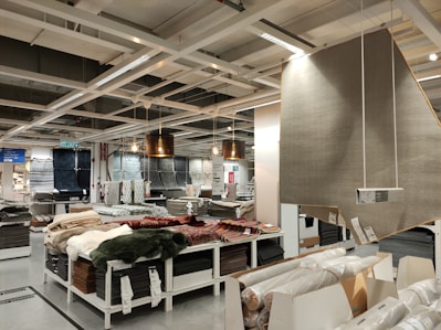 A retail store interior featuring a large selection of rolled-up carpets and rugs displayed on shelves and tables. Several hanging lamps provide lighting to the area, and a variety of carpet styles and colors are visible. The shelving units are neatly organized with packaged flooring products, and a large sign displaying product information is visible.