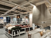 A retail store interior featuring a large selection of rolled-up carpets and rugs displayed on shelves and tables. Several hanging lamps provide lighting to the area, and a variety of carpet styles and colors are visible. The shelving units are neatly organized with packaged flooring products, and a large sign displaying product information is visible.