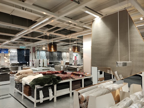 A retail store interior featuring a large selection of rolled-up carpets and rugs displayed on shelves and tables. Several hanging lamps provide lighting to the area, and a variety of carpet styles and colors are visible. The shelving units are neatly organized with packaged flooring products, and a large sign displaying product information is visible.