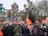 A large group of people gathered in front of a historic building in a public space. Many individuals are holding red flags with white logos and text. People are wearing winter clothing, indicating a cold day. The scene is set against a backdrop of leafless trees that are pruned in a unique manner, and road signs are visible, showing directions. The mood suggests a protest or public demonstration.