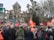 A large group of people gathered in front of a historic building in a public space. Many individuals are holding red flags with white logos and text. People are wearing winter clothing, indicating a cold day. The scene is set against a backdrop of leafless trees that are pruned in a unique manner, and road signs are visible, showing directions. The mood suggests a protest or public demonstration.