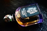 A luxurious perfume bottle with a metallic cap and amber-colored liquid, featuring an ornate and stylish label displaying intricate designs. It is placed on a dark, reflective surface.