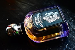 A luxurious perfume bottle with a metallic cap and amber-colored liquid, featuring an ornate and stylish label displaying intricate designs. It is placed on a dark, reflective surface.