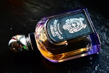 A luxurious perfume bottle with a metallic cap and amber-colored liquid, featuring an ornate and stylish label displaying intricate designs. It is placed on a dark, reflective surface.