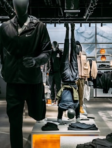 Mannequins display athletic apparel in a retail setting, with one mannequin doing a headstand. The background features shelves and clothing, while the foreground includes folded garments. Bright lighting highlights the store setup.