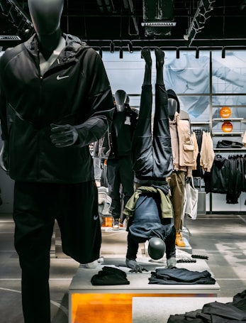 Mannequins display athletic apparel in a retail setting, with one mannequin doing a headstand. The background features shelves and clothing, while the foreground includes folded garments. Bright lighting highlights the store setup.