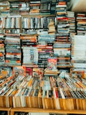A tightly packed collection of books fills bookshelves and covers a table, with stacks upon stacks creating a complex and dense formation. Various titles of different genres and sizes are visible, contributing to an atmosphere of organized chaos.