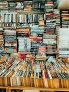 A tightly packed collection of books fills bookshelves and covers a table, with stacks upon stacks creating a complex and dense formation. Various titles of different genres and sizes are visible, contributing to an atmosphere of organized chaos.