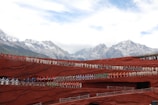 A large ensemble of people dressed in vibrant traditional costumes stand in rows on terraced red structures against a backdrop of majestic snow-capped mountains and a partly cloudy sky.