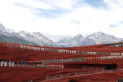A large ensemble of people dressed in vibrant traditional costumes stand in rows on terraced red structures against a backdrop of majestic snow-capped mountains and a partly cloudy sky.