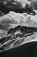 A dramatic black and white photograph of a mountain range with snow-capped peaks under a cloudy sky. The clouds are thick and create a moody contrast with the dark mountains.