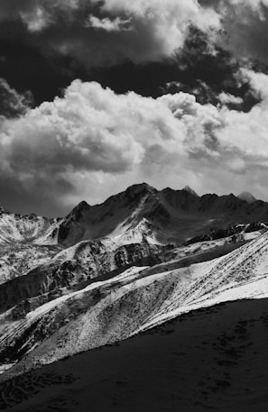 A dramatic black and white photograph of a mountain range with snow-capped peaks under a cloudy sky. The clouds are thick and create a moody contrast with the dark mountains.