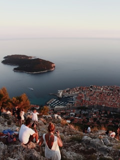 A scenic view showcases people sitting and taking photos at a rocky vantage point, overlooking a historic coastal town with a marina visible. The expansive sea extends outward, with a small lush island nearby.