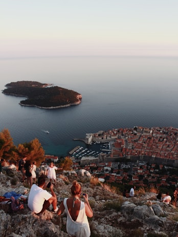 A scenic view showcases people sitting and taking photos at a rocky vantage point, overlooking a historic coastal town with a marina visible. The expansive sea extends outward, with a small lush island nearby.