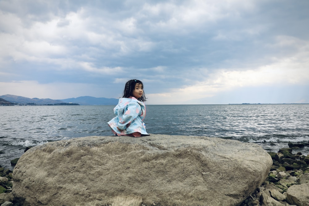 a little girl sitting on a rock by the water