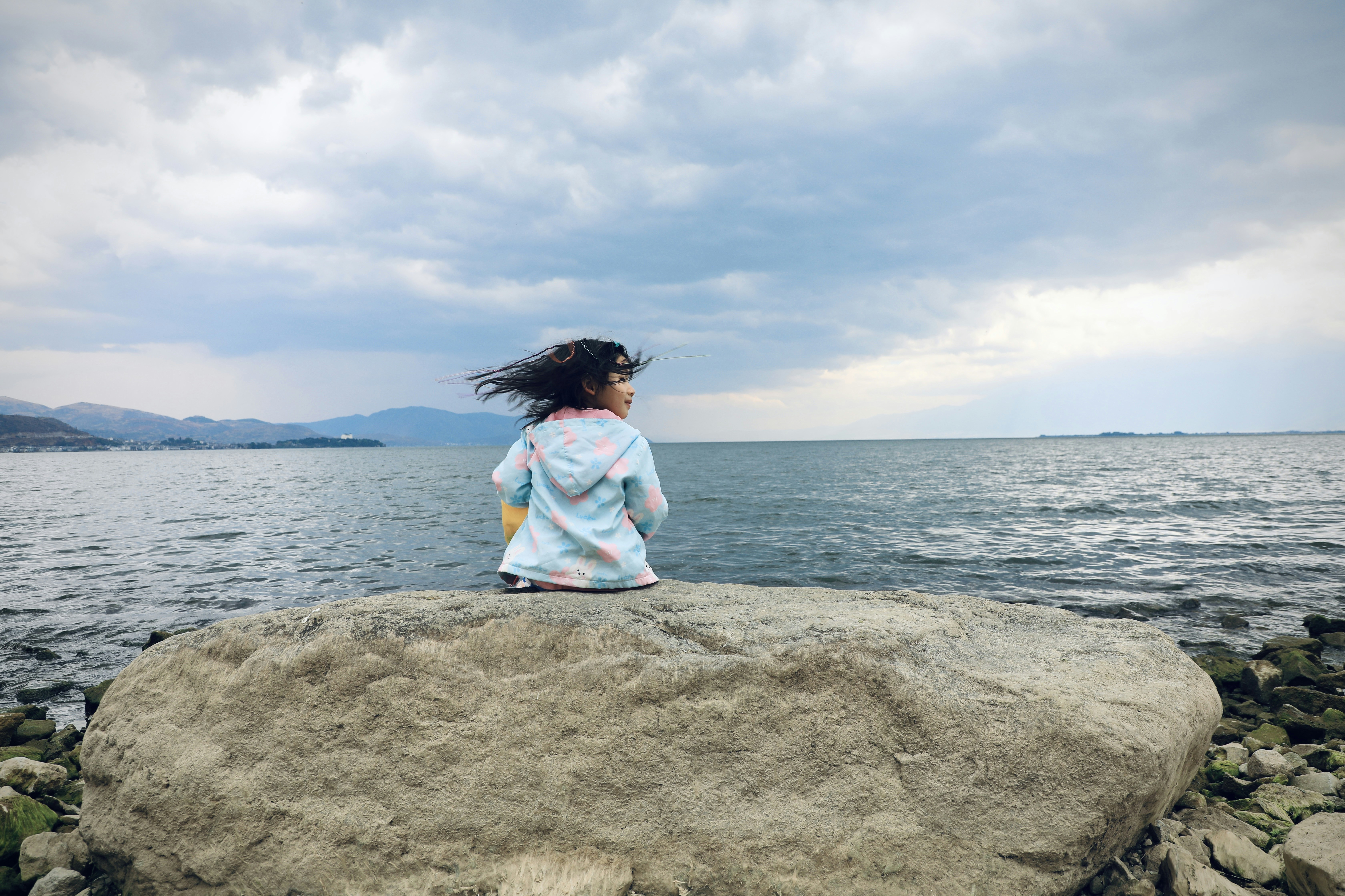 A girl sitting on a rock looking out at the water photo – Free Lake ...