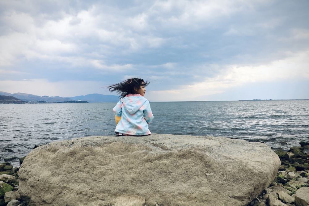 a girl sitting on a rock looking out at the water