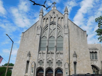 A large stone church with intricate Gothic architectural details and a large central window. The building features ornate carvings and spires, conveying a historical and religious atmosphere. A flagpole with the American flag is seen to the left, and trees surround the building under a partly cloudy blue sky.