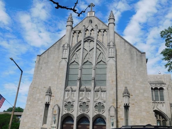 A large stone church with intricate Gothic architectural details and a large central window. The building features ornate carvings and spires, conveying a historical and religious atmosphere. A flagpole with the American flag is seen to the left, and trees surround the building under a partly cloudy blue sky.