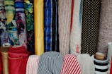 Various colorful rolls of fabric are stacked vertically, showcasing a range of patterns including floral, solid colors, stripes, and polka dots. The fabrics have different textures and colors, from bright yellow and deep reds to calming blues and muted earth tones.