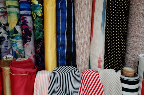 Various colorful rolls of fabric are stacked vertically, showcasing a range of patterns including floral, solid colors, stripes, and polka dots. The fabrics have different textures and colors, from bright yellow and deep reds to calming blues and muted earth tones.