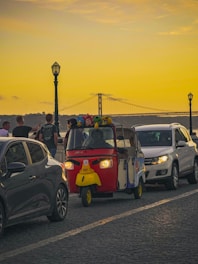 A vibrant tuk-tuk decorated with colorful plush toys is driving alongside cars on a cobblestone road. The background features a large bridge under a yellow-orange sunset sky, creating a calm and lively atmosphere. A few people are visible near the tuk-tuk, indicating a casual street scene.