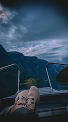 A serene landscape featuring a boat ride through a narrow fjord with tall, lush green mountains on either side. The sky is overcast, creating a moody atmosphere. The foreground shows a person's legs and feet resting on the boat deck, emphasizing a relaxed, leisurely vibe.