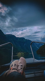 A serene landscape featuring a boat ride through a narrow fjord with tall, lush green mountains on either side. The sky is overcast, creating a moody atmosphere. The foreground shows a person's legs and feet resting on the boat deck, emphasizing a relaxed, leisurely vibe.
