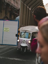 A white tuk-tuk with a patterned front is driving through a construction area, next to a corrugated metal fence with a sign written in Portuguese. The foreground is blurred with a person on the right side of the image.