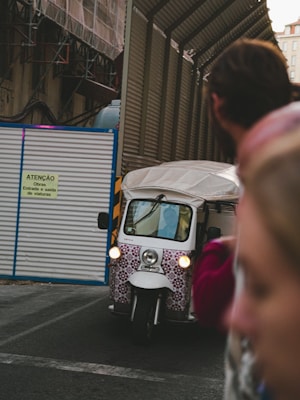 A white tuk-tuk with a patterned front is driving through a construction area, next to a corrugated metal fence with a sign written in Portuguese. The foreground is blurred with a person on the right side of the image.