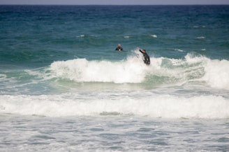 Two surfers in black wetsuits are riding the waves in the ocean, with one surfer more prominently visible maneuvering through the surf. The sea is a mix of turquoise and deep blue, creating dynamic waves under a clear sky.