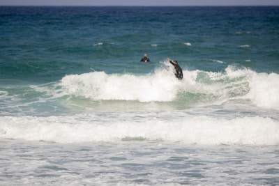 Two surfers in black wetsuits are riding the waves in the ocean, with one surfer more prominently visible maneuvering through the surf. The sea is a mix of turquoise and deep blue, creating dynamic waves under a clear sky.