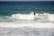 Two surfers in black wetsuits are riding the waves in the ocean, with one surfer more prominently visible maneuvering through the surf. The sea is a mix of turquoise and deep blue, creating dynamic waves under a clear sky.