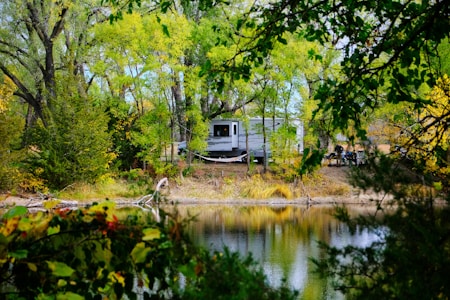 A recreational vehicle is parked amidst a lush forest by a calm, reflective lake. The area is dense with green foliage, and the trees are full and vibrant. The mirrored surface of the water beautifully reflects the surrounding trees and sky. A few people are gathered near the RV, possibly enjoying a camping trip.