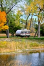 A small camper is parked near a tranquil lake, surrounded by tall trees with leaves transitioning into autumn colors. A picnic table is situated close to the camper, and the ground is covered with grass and fallen leaves. The scene is peaceful and natural, with the water reflecting the trees and sky.