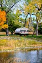 A small camper is parked near a tranquil lake, surrounded by tall trees with leaves transitioning into autumn colors. A picnic table is situated close to the camper, and the ground is covered with grass and fallen leaves. The scene is peaceful and natural, with the water reflecting the trees and sky.
