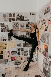 A woman with tattoos strikes a dynamic pose in a room covered with newspapers and magazine clippings. The clippings feature various images and text, adding a collage-like backdrop. She wears a black tank top and pants, with one leg raised against the wall. Her expression is playful and confident.