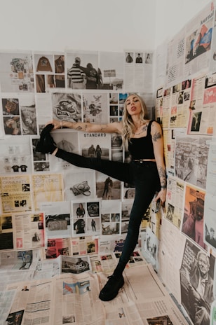 A woman with tattoos strikes a dynamic pose in a room covered with newspapers and magazine clippings. The clippings feature various images and text, adding a collage-like backdrop. She wears a black tank top and pants, with one leg raised against the wall. Her expression is playful and confident.