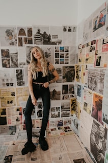 A woman with long blonde hair and tattoos stands in a room covered with newspaper clippings on the walls and floor. She is wearing a black sleeveless top, dark jeans, and black shoes. The clippings feature various images and text in different styles, showcasing a vintage and eclectic aesthetic.
